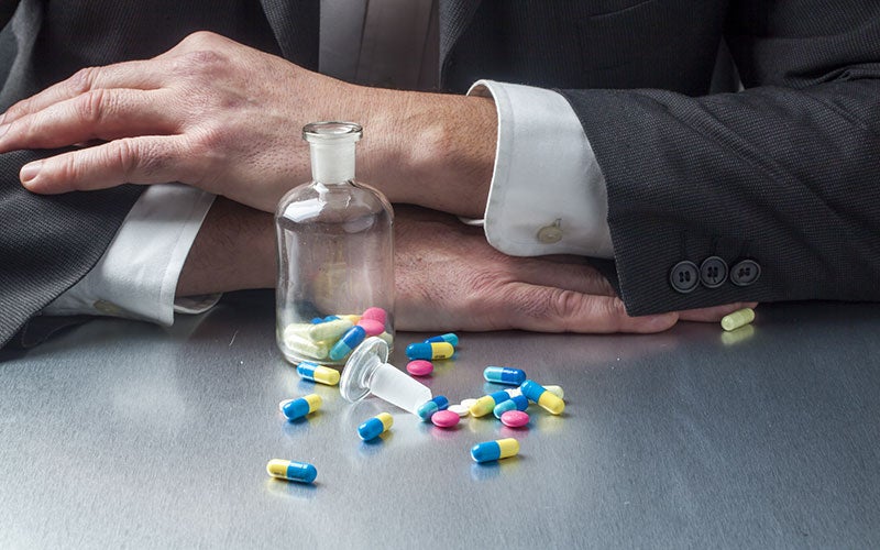Pill bottle open with pills around it on desk, man in suit arms crossed in front
