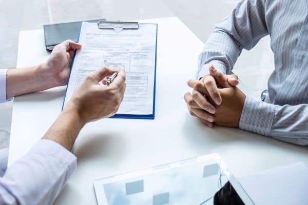 A close up of a doctor explaining what is on a clipboard to a patient with hands clasped at the desk