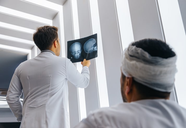 A man with head bandages sits in a hospital room with a doctor as they view x-rays of the man's head