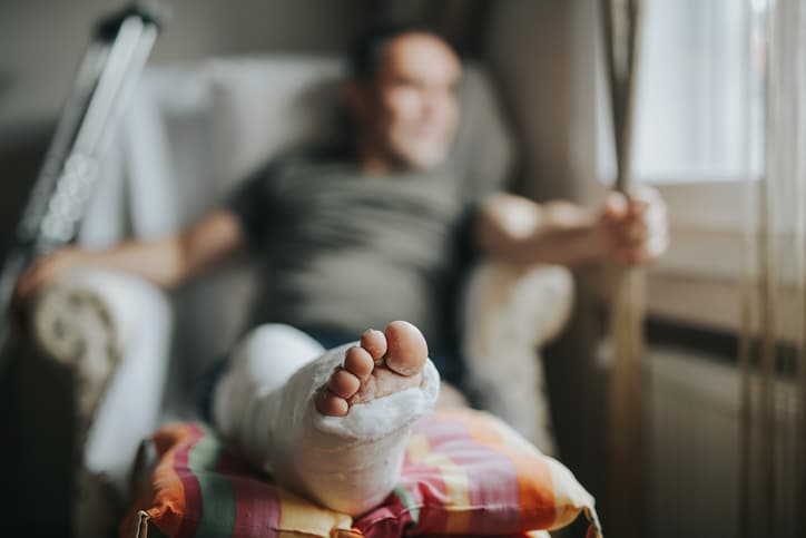 A man sitting on a recliner at home is blurred out in the background but his leg in a cast on a foot stool is in focus