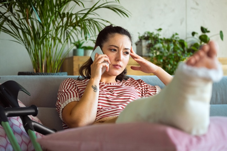 Worried young woman with a broken leg in plaster cast sitting on a sofa at home and listening to someone on the phone