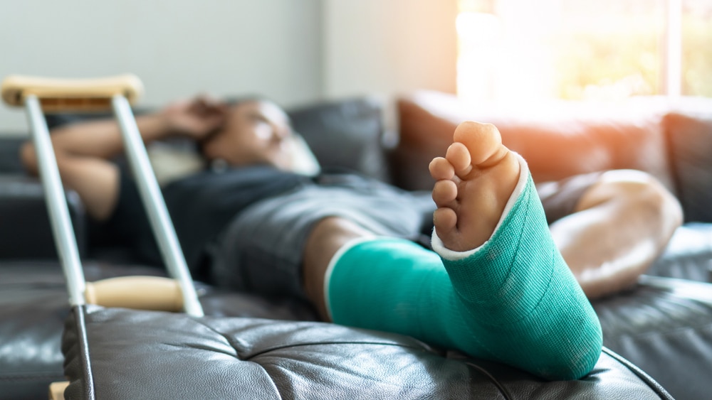 A man lays at home on the couch with a broken leg and a crutch nearby