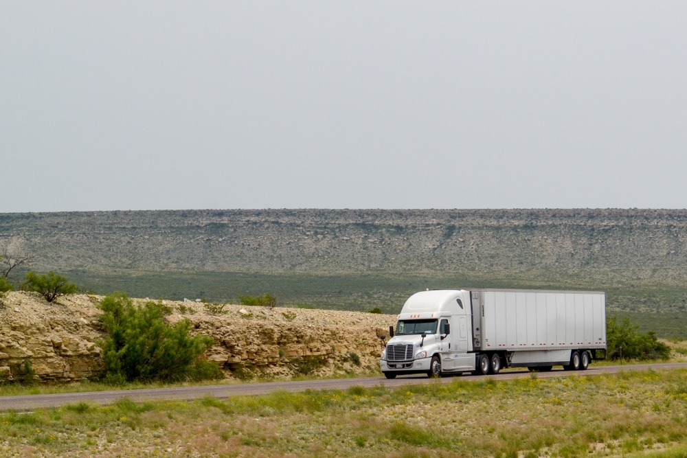 A white semi truck travels down the highway in a desert landscape with bluffs in the background